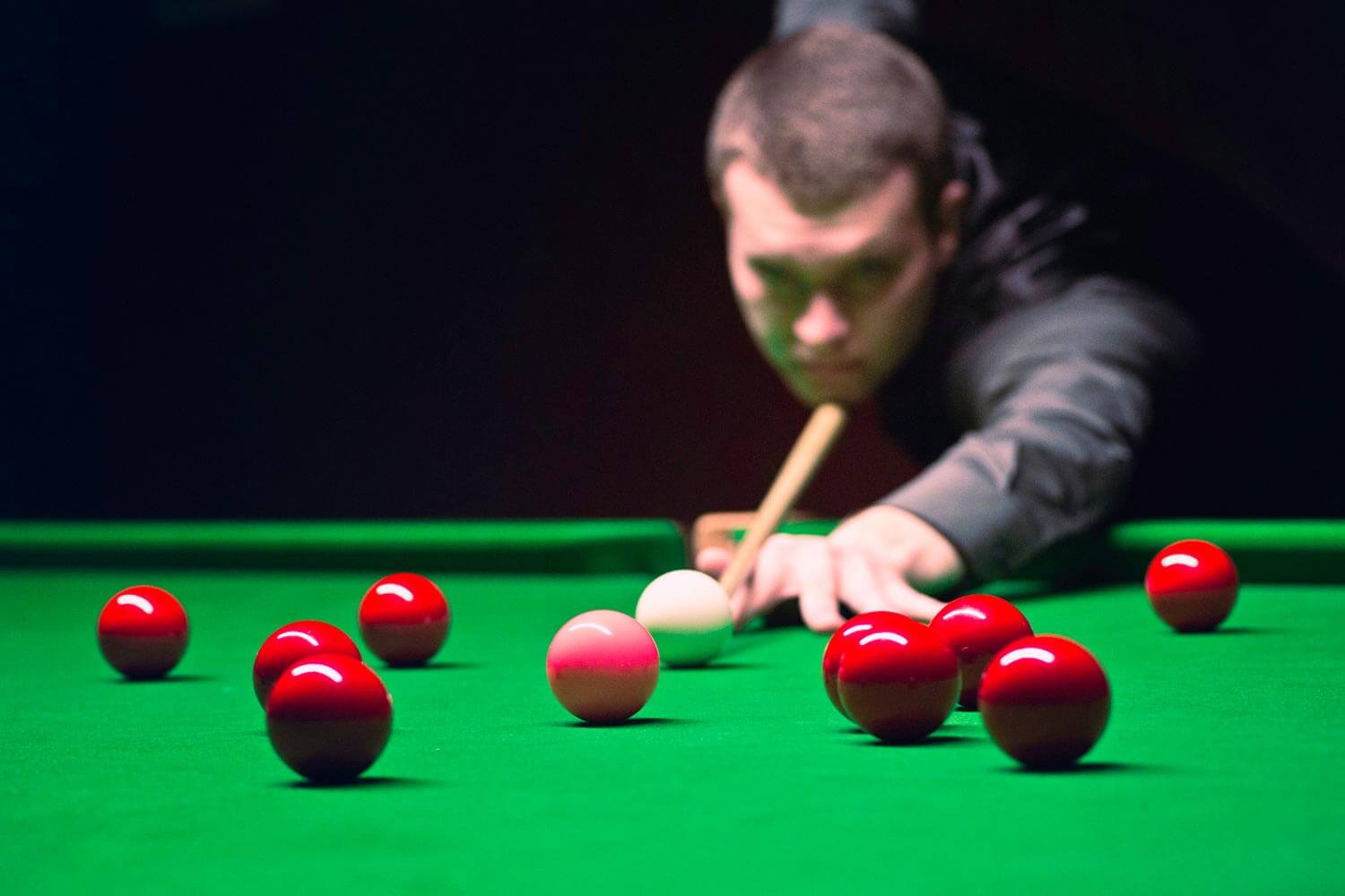 A focused snooker player lining up a shot with a cue stick on a green baize snooker table, with red snooker balls in the foreground A focused snooker player lining up a shot with a cue stick on a green baize snooker table, with red snooker balls in the foreground